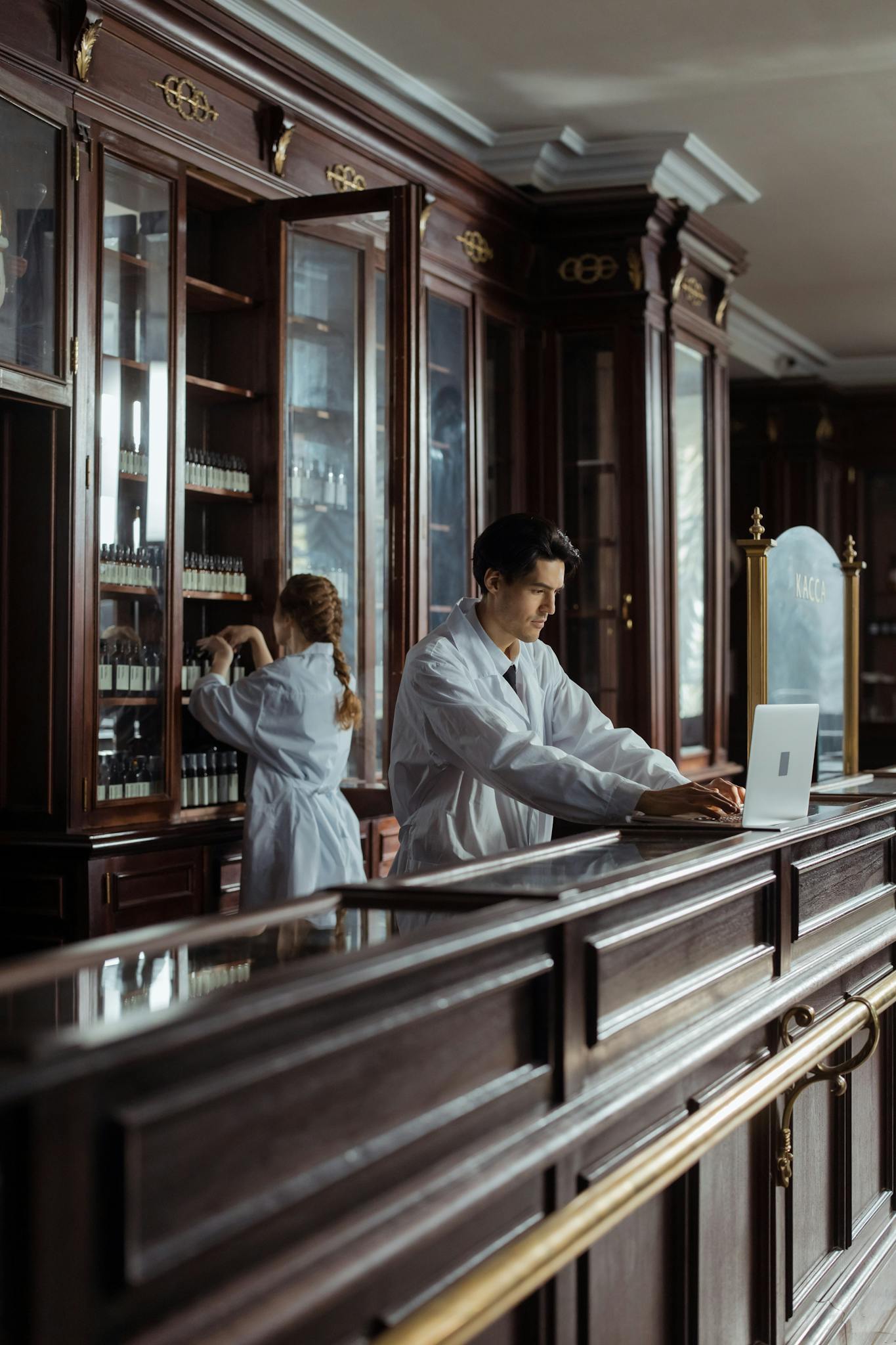 Pharmacists in a traditional pharmacy setting with wooden cabinetry and a laptop.